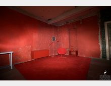 Ein leerer Raum mit roten Wänden, Teppichboden und minimaler Einrichtung, darunter ein roter Sessel, eine Stehlampe, ein Sideboard und ein kleiner Beistelltisch mit Vase. Die Wände zeigen abgeblätterte Farbe und freiliegende Ziegel, was auf einen vernachlässigten oder unrenovierten Zustand hinweist.