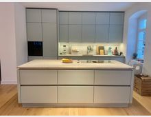 Moderne, minimalistische Küche mit hellgrauen, grifflosen Schrankfronten und einer großen Kücheninsel mit Steinplatte und integriertem Kochfeld. An der Rückwand verlaufen Hängeschränke und Arbeitszeile mit Spüle und Unterbauleuchte; rechts sorgt ein hohes Fenster in einer Nische für Tageslicht.