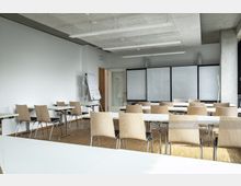 Moderner Seminarraum mit hellen Holzböden, minimalistischen Holzstühlen, weißen rechteckigen Tischen und großen schwarzen Whiteboards im Hintergrund. An einer Wand steht ein Flipchart, und Tageslicht fällt durch ein Fenster auf der rechten Seite ein.