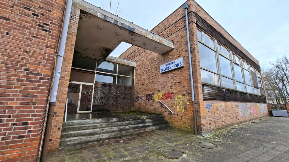 Eingangsbereich eines Backsteingebäudes mit Treppenaufgang, Glasfront und auskragendem Betonvordach; an der Fassade hängt ein Schild „Theater-Café“. Große Fensterreihen und Graffiti an den Wänden prägen die Außenansicht, davor liegt ein gepflasterter Vorplatz mit Bäumen im Hintergrund.