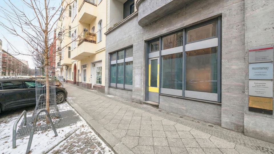 Straßenszene mit breitem Gehweg vor einem grauen Gebäude mit großer Schaufensterfront und Eingangstür sowie einem Schild an der Fassade. Entlang der Straße stehen parkende Autos und ein kahler Straßenbaum, am Boden liegen stellenweise Schnee und Pflastersteine.