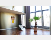 Heller Wohnraum mit Holzboden, einer großen Fensterfront mit bodentiefen Glastüren und grauen Vorhängen. Im Raum stehen ein Liegestuhl mit Fellbezug und eine große Zimmerpalme in einem Terrakottatopf; an der goldfarbenen Wand hängt ein Bild einer Frau in einem gelben Kleid.