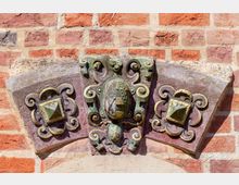 Nahaufnahme eines dekorativen Reliefs aus Stein und Metall auf einer Backsteinfassade. Das Relief zeigt filigrane Verzierungen, ein zentral platziertes Wappen und flankierende geometrische Ornamente mit goldfarbenen Elementen.