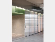Innenraum mit modernen industriellen Elementen, einschließlich transluzenter, verschiebbarer Metallpaneele, einer rohen Betonwand und Deckenverkleidungen aus Metall. Im oberen Bereich befindet sich ein beleuchtetes Konstrukt aus grünem Glas.