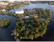 Luftaufnahme einer von Wasser umgebenen bewaldeten Insel mit Gebäuden, darunter mehrere Backsteinbauten und ein weißes Hochgebäude. Die Insel ist von Bootsanlegestellen und einer grünen Umgebung geprägt, während im Hintergrund Stadtgebiete und weitere Gewässer sichtbar sind.