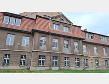 Alt-Text: Außenansicht eines mehrstöckigen, historischen Gebäudes mit brauner Fassade, weißen Fenstern und roten Ziegeldächern. Das Erdgeschoss zeigt große Gitterfenster, während die oberen Stockwerke kleinere Fenster und ein Giebeldach haben.