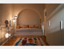 Kleines, gemütlich eingerichtetes Zimmer mit heller Wandfarbe; im Zentrum eine in die Wand integrierte Sitznische mit beigen Kissen, daneben ein Regal mit Deko-Elementen und eine moderne Stehleuchte. Im Vordergrund ein bunter Teppich auf Holzboden, rechts ein großer weißer Einbauschrank.