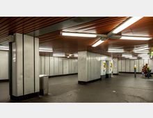 Unterirdischer U-Bahn-Bereich mit metallischen Wandverkleidungen, einer dunkelgrauen Bodenfläche und einer Decke aus Holzpaneelen mit deckenmontierten Leuchtstofflampen. Mehrere Fahrkartenautomaten sind in einer Nische sichtbar, und im Hintergrund stehen Menschen mit einem Kinderwagen.