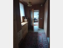 Ein schmaler Flur mit Holzvertäfelung an Wänden und Decke, ausgestattet mit einem orientalischen Teppich und einem hölzernen Sideboard. Eine geöffnete Zimmertür führt zu einem angrenzenden Raum mit einem Esstisch und Stühlen, beleuchtet durch ein Fenster mit weißen Vorhängen.