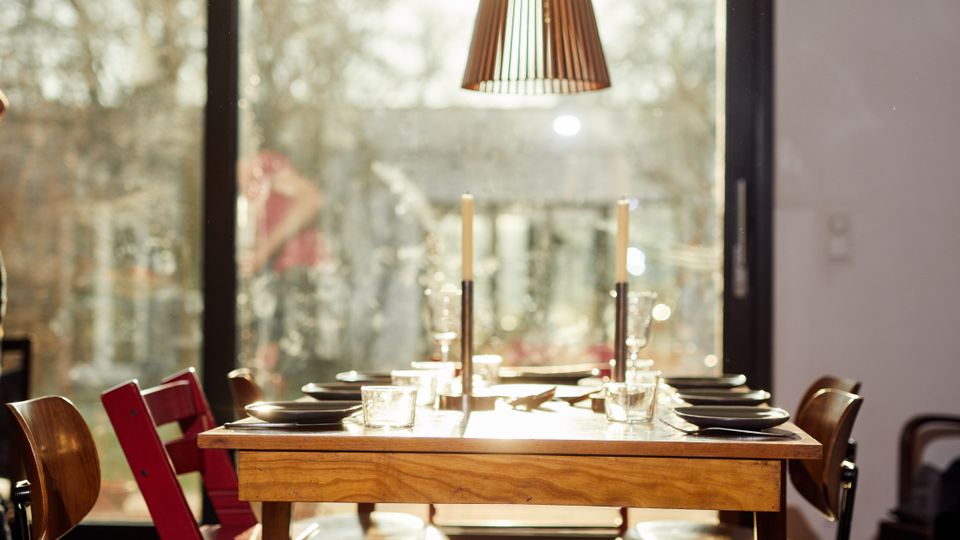A dining area with a wooden table set for a meal, surrounded by wooden chairs, and illuminated by sunlight streaming through a large glass sliding door. A modern, cone-shaped pendant light hangs above, and the outdoor scenery is partially visible.
