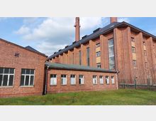 Backsteinindustriegebäude mit einer flachen Anbauhalle und hohen, vertikalen Fenstern im Haupthaus. Im Hintergrund ragen zwei lange Schornsteine in den Himmel, umgeben von einer Grünfläche im Vordergrund.