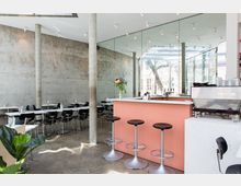 Modernes Café im minimalistischen Design mit Sichtbetonwänden, einer Theke in Rosatönen, schwarzen Barhockern und schwarzen Stühlen an weißen Tischen. Große Fensterfronten und Spiegel reflektieren das Tageslicht, während Pflanzen als Dekoration dienen.