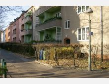 Mehrstöckige Wohngebäude in einer Wohnstraße mit Balkonen, wobei die Fassaden in verschiedenen Pastelltönen gestrichen sind. Im Vordergrund sind ein Gehweg, eine niedrige Hecke mit Herbstlaub und eine Straßenlaterne zu sehen.