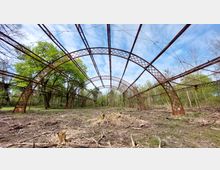 Rostige Stahlkonstruktion eines ehemaligen Gewächshauses oder Gebäudes in einer bewaldeten Umgebung, mit kahlem Boden im Vordergrund und grünen Bäumen im Hintergrund unter einem bewölkten blauen Himmel.