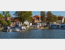 Riverside marina with several small boats and a larger cabin cruiser moored along wooden docks on calm water. Behind the docks are leafy trees and a row of low buildings with red-tiled roofs and light-colored facades.