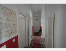 Ein schmaler Flur in einem Wohnhaus mit weißen Wänden, teilweise rot gestrichen. Links hängt ein Poster mit Fahrradillustrationen, rechts steht ein zusammengeklappter Regenschirm. Ein schmaler Teppich in Rot- und Blautönen liegt auf dem Holzboden, und am Ende des Flurs befindet sich eine weiße Eingangstür.