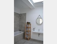 Modernes Badezimmer mit grauen Fliesen und einer Badewanne auf der linken Seite. Rechts steht ein Holzregal mit Körben neben einem Waschbecken mit Spiegel darüber; natürliche Beleuchtung durch ein Oberlicht.