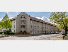 Mehrstöckiges Wohngebäude mit heller Putzfassade und roten Ziegeldächern an einer urbanen Straße, flankiert von Bäumen. Am Straßenrand parken mehrere Autos, und ein Gehweg verläuft entlang der Häuserfront.