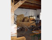 Innenansicht eines rustikalen Raumes mit Holzbalkendecke und weiß verputzten Wänden. Im Mittelpunkt steht ein hölzerner Handwagen, beladen mit Heu und Holz, daneben befinden sich Holzbänke und ein Tisch auf einem Holzboden.