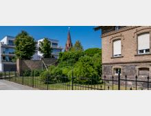 Ein Stadtbild mit einem modernen Wohngebäude links, einer alten Backsteinfassade rechts und einem hohen Kirchturm aus rotem Stein im Hintergrund. Im Vordergrund sind ein Zaun, eine Rasenfläche und dichter Bewuchs sichtbar.