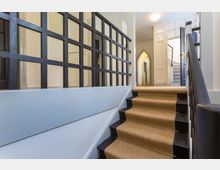 Treppenhaus mit schwarzen Holzgeländern und einem mit Teppich verkleideten Treppenlauf. Oben befinden sich weiße Wände, eine Tür mit Glaselementen und ein gewölbter Durchgang.