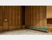 Holzvertäfelter Innenraum mit hohen Wänden und akustischen Elementen, bestehend aus Holzgitterpaneelen in geometrischem Design. Im unteren Bereich befindet sich eine lange grüne Polsterbank, links eine Holztür mit einem grünen Notausgangsschild, und der Boden ist mit Fischgrätparkett ausgelegt.