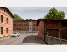 Straßenszene in einem historischen Viertel mit Backsteingebäuden, darunter ein Wohnhaus im Hintergrund mit verzierten Fenstern und ein Nebengebäude rechts mit Holztüren. Im Vordergrund ein geparktes Auto und ein Gehweg mit Metallzaun, umgeben von einzelnen Bäumen.