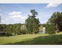 Ein weitläufiger Park mit einer grünen Wiese, vereinzelt großen Bäumen und einem See im Hintergrund. Auf der gegenüberliegenden Uferseite ist ein Gebäude mit heller Fassade und dunklem Dach zu sehen, umgeben von dichter, bewaldeter Vegetation.