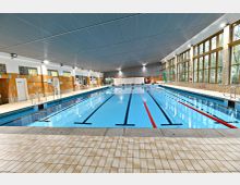 Innenansicht eines Hallenbads mit einem großen rechteckigen Schwimmbecken, das in mehrere Bahnen unterteilt ist. Die Wände bestehen aus gelblichen Fliesen, großen Fenstern und Glaselementen; die Decke ist aus grauem Metall, der Boden ist mit hellen Fliesen ausgelegt.