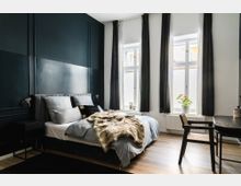 Modernes Schlafzimmer mit dunkelgrüner Wand und einem Doppelbett, das mit grauer Bettwäsche und einer beigen Felldecke ausgestattet ist. Drei hohe Fenster mit schwarzen Vorhängen lassen Tageslicht herein, ein Schreibtisch mit Stuhl steht rechts, ein kleiner Blumentopf auf der Fensterbank.