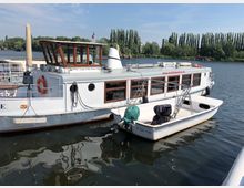 Ein Bootsanleger mit einem größeren weißen Ausflugsboot mit Holzfenstern und einem kleineren Motorboot im Vordergrund. Im Hintergrund ist ein ruhiger Fluss mit Bäumen und grasbewachsenen Ufern zu sehen.