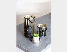 Zwei schwarze, zylindrische Laternen mit goldfarbener Innenbeschichtung und Tragegriffen stehen auf einer grauen Oberfläche, daneben ein kleiner weißer Blumentopf mit einer grünen Sukkulente.