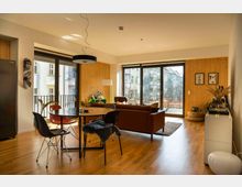 Helles Wohnzimmer mit Holzboden, einer braunen Ledercouch und einem runden Esstisch mit schwarzen und roten Stühlen. Große Fensterfronten und Glastüren bieten Blick auf einen Balkon und dahinterliegende Wohngebäude; die Wände sind teilweise mit hellem Holz verkleidet.