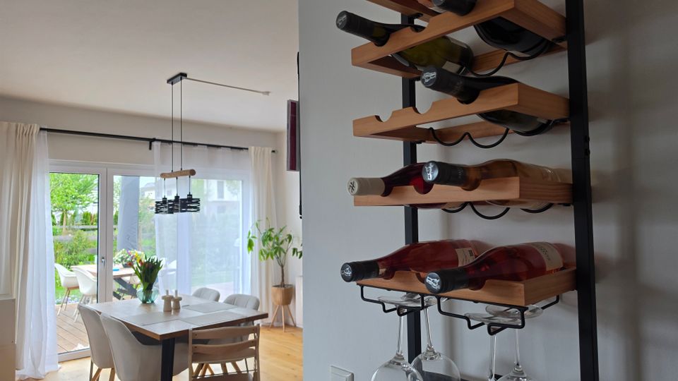 Moderner Essbereich mit Holzfußboden, Esstisch und Stühlen vor großen Schiebetüren mit weißen Vorhängen, die auf eine Terrasse und einen Garten führen. An der Wand im Vordergrund hängt ein schwarzes Metall-Weinregal mit Holzablagen für Flaschen und hängenden Weingläsern.