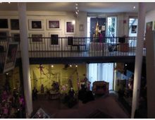Zweistöckiger Museumsraum mit einer Galerieebene, dekoriert mit Puppen, Figuren und Kunstwerken an den Wänden. Unten befinden sich Ausstellungsvitrinen mit Puppenthemen, während oben Gemälde hängen und Möbel ausgestellt sind, teils vor großen Fenstern mit Tageslicht.