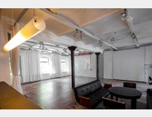 Großzügiger, industrieller Innenraum mit weißen Wänden und Decke sowie sichtbaren Metallträgern. Der Raum verfügt über bodentiefe Fenster mit weißen Vorhängen, eine schwarze Ledersitzgruppe und eine moderne Hängelampe.