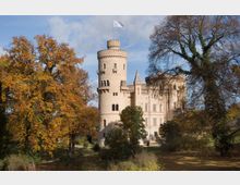 Eine historische Schlossanlage mit heller Fassade und einem runden Turm, der von einer weißen Fahne bekrönt ist. Das Schloss ist von Herbstbäumen und einer gepflegten Gartenlandschaft umgeben.