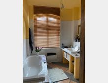 Badezimmer mit Badewanne links, einem großen Fenster mit Holzjalousien in der Mitte und einem Waschtisch mit gelbem Sockel rechts. Die Wände sind teilweise gelb gestrichen und teils weiß gefliest, der Boden ist mit braunen und grauen Fliesen versehen.