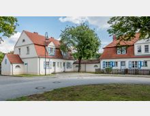 Zwei weiß verputzte Wohnhäuser mit roten Ziegeldächern und blauen Fensterläden stehen an einer gepflasterten Straße, getrennt durch einen Baum und einen kleinen Zaun. Die Umgebung ist von Rasenflächen und vereinzelten Bäumen geprägt.