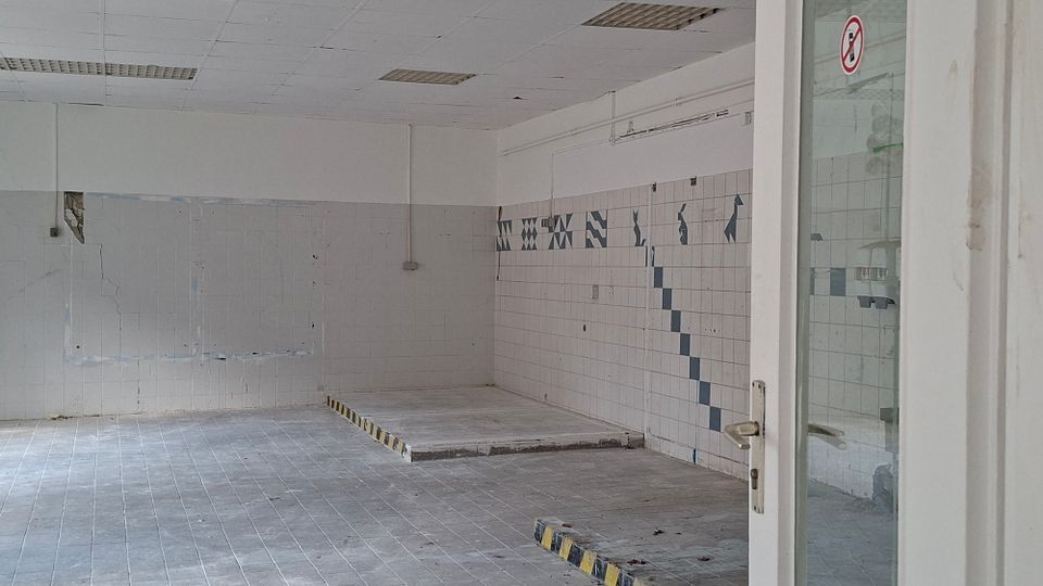 Leerer, gefliester Innenraum eines ehemaligen Industrie- oder Küchenbereichs mit erhöhter Plattform und Wänden aus weißen Fliesen mit blauen Mustern. Abgehängte Decke mit Leuchtstofflampen und eine Glastür mit Verbotsschild am rechten Bildrand.