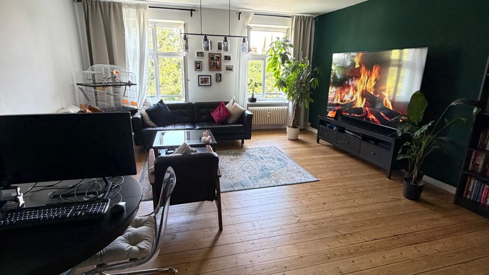 Helles Wohnzimmer mit breiten Holz-Dielen, dunkler Akzentwand und großem Flachbildfernseher auf einem Lowboard. Vor zwei großen Fenstern mit Vorhängen stehen Sofa und Couchtisch auf einem Teppich; links ist ein Arbeitsplatz mit Schreibtisch, Monitoren und Stuhl zu sehen.
