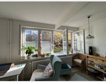 Helles Wohnzimmer mit großen Fenstern, durch die ein Garten mit herbstlichen Bäumen sichtbar ist. Im Raum stehen ein grüner Sessel, ein kleiner Tisch mit Laptop, eine Holz-Kommode und Zimmerpflanzen auf der Fensterbank.