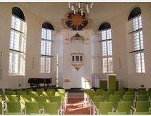 Lichterfüllter Innenraum einer Kirche mit hohen, rundbogigen Fenstern, einem weißen Kanzelaltar mit goldenen Verzierungen und einer dekorativen goldenen Sonne oberhalb. Der Raum ist mit grünen Stühlen in Reihen bestuhlt, neben dem Altar steht ein Flügel.