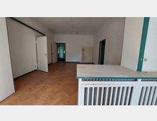 Ein leerer Raum mit hellen Wänden, einem Holzparkettboden und zwei offenen Türen, die in verschiedene Bereiche führen. Im Vordergrund befindet sich eine weiße Heizkörperverkleidung, und der Raum ist von einem schlichten, funktionalen Stil geprägt.