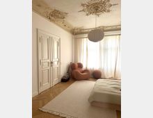 Helles Schlafzimmer mit Stuckdecke, weißen Wänden und Holzboden in Fischgrätenmuster. Ein großer Teppich, ein minimalistisches Bett und ein gestreifter, organisch geformter Sessel prägen den Raum, der von bodenlangen, lichtdurchlässigen Vorhängen erhellt wird.