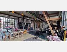 Großzügiger, modern eingerichteter Raum mit Loft-Charakter, erkennbar an hohen Decken, großen Fenstern und sichtbaren Stahlträgern. Im Vordergrund ein Esstisch mit Stühlen, im Hintergrund eine offene Küche, ein Billardtisch, ein Klavier und eine Metalltreppe zu einer Galerieebene.