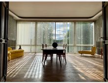 Großzügiges Wohnzimmer mit großen Fensterfronten, die mit vertikalen Lamellenjalousien versehen sind. Der Raum ist minimalistisch möbliert mit einem Holztisch und Stühlen in der Mitte, einem gelben Sofa links und einem gelben Sessel rechts; die Aussicht zeigt Bäume und einen sonnigen Himmel.