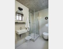 Modern bathroom with white tiled walls, a concrete ceiling, and a glass-enclosed corner shower. A wall-mounted sink beneath an angular mirror, a hanging towel, and a wall-hung toilet sit on a smooth light-gray floor.
