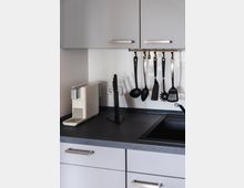 Graue moderne Küchenzeile mit Hängeschränken, einer dunklen Arbeitsplatte, einem weißen Kaffeeautomaten und einer schwarzen Spüle. Küchenutensilien wie Schöpfkellen und Pfannenwender hängen an einer Stange über der Arbeitsfläche.