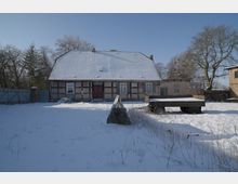 Verschneiter Hof vor einem niedrigen Fachwerkhaus mit großem, schneebedecktem Satteldach und mittiger Eingangstür. Rechts steht ein flacher Anhänger im Schnee; im Hintergrund sind kahle Bäume und Nebengebäude zu sehen.